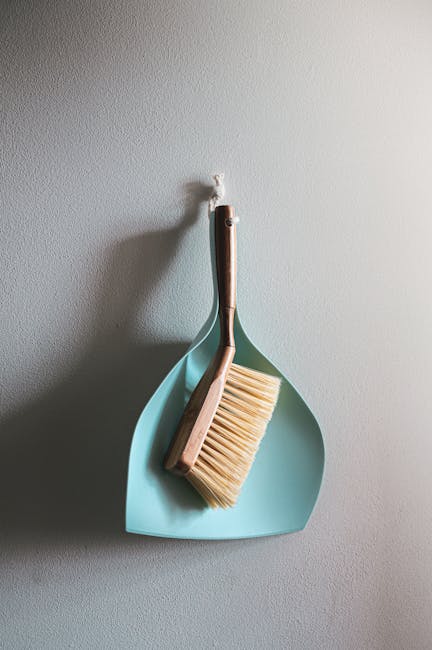 A light blue dustpan and a wooden-handled brush with beige bristles hanging on a white wall in a clean and well-lit indoor space, illustrating surface cleaning tools used in domestic cleaning by Carpe